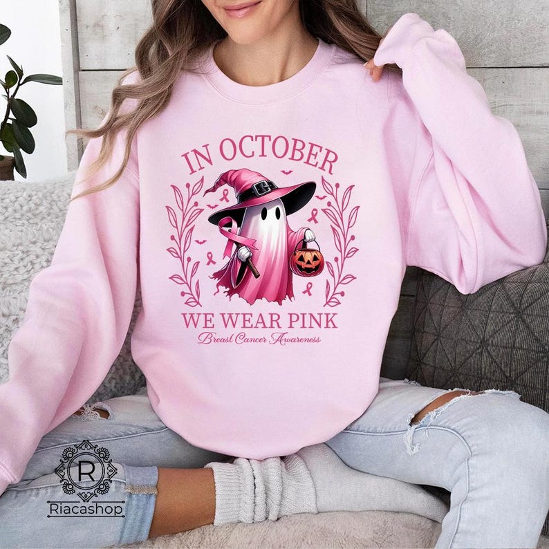 May include: A light pink sweatshirt with a graphic featuring a ghost wearing a witch hat and holding a pumpkin. The text reads "IN OCTOBER WE WEAR PINK Breast Cancer Awareness".