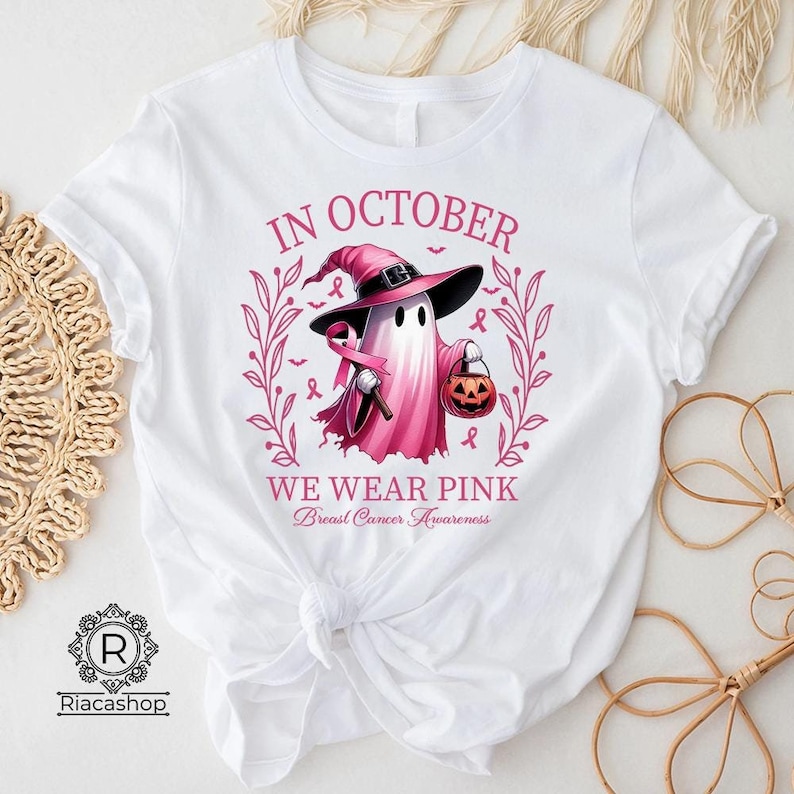May include: White t-shirt with a graphic of a pink ghost wearing a witch hat and holding a pink ribbon and a jack-o'-lantern. The text reads "IN OCTOBER WE WEAR PINK Breast Cancer Awareness".