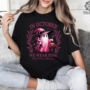 May include: Black t-shirt with a pink graphic featuring a ghost wearing a witch hat and holding a pumpkin. The text reads "In October We Wear Pink" with "Breast Cancer Awareness" below.