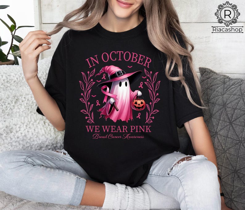 May include: Black t-shirt with a pink graphic featuring a ghost wearing a witch hat and holding a pumpkin. The text reads "In October We Wear Pink" with "Breast Cancer Awareness" below.