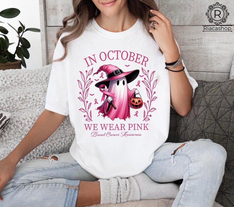 May include: White t-shirt with a pink and black graphic. The graphic features a ghost wearing a witch hat and holding a jack-o'-lantern, with the text "IN OCTOBER WE WEAR PINK Breast Cancer Awareness".