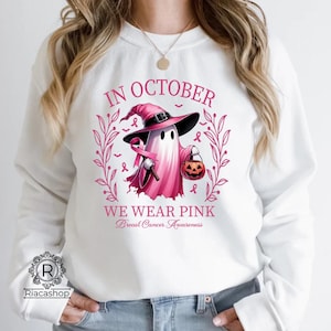 May include: White sweatshirt with a pink graphic that says "IN OCTOBER WE WEAR PINK Breast Cancer Awareness". The graphic features a ghost wearing a witch hat and holding a pink ribbon and a jack-o'-lantern.