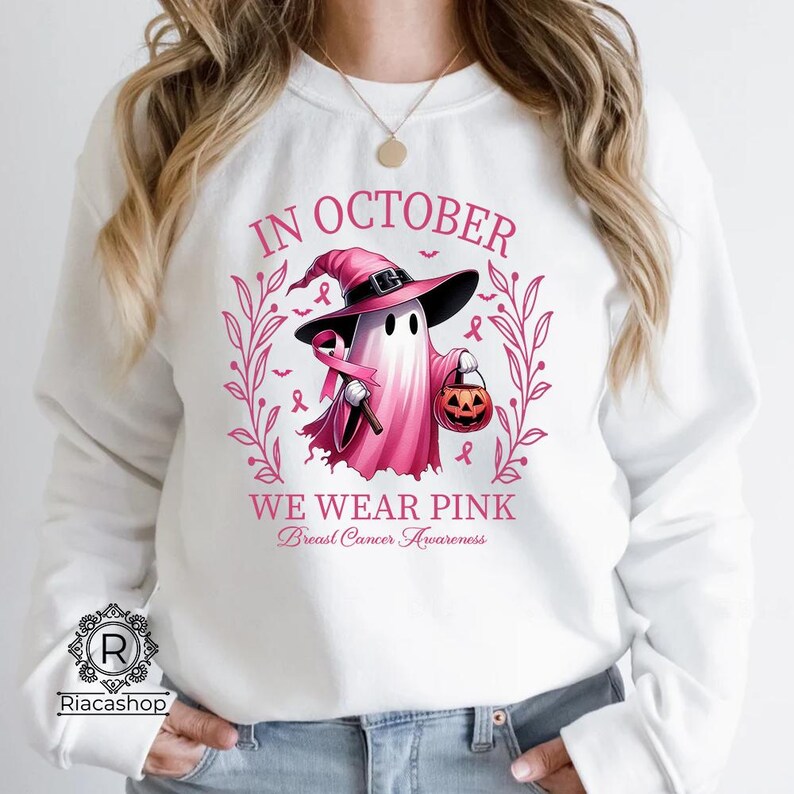 May include: White sweatshirt with a pink graphic that says "IN OCTOBER WE WEAR PINK Breast Cancer Awareness". The graphic features a ghost wearing a witch hat and holding a pink ribbon and a jack-o'-lantern.