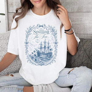 May include: A white short-sleeve t-shirt featuring a blue graphic print of a sailing ship with the text Spilling The Tea and Since 1775. The design is enclosed in an ornate floral oval frame with bows at the top and sides, depicting a historical scene.