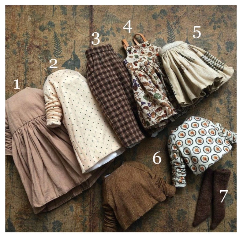 May include: A collection of doll clothes in various styles and patterns. Includes a tan dress, a cream-colored jacket with black polka dots, brown plaid pants, a floral print dress, a tan skirt, a patterned jacket, and brown leggings.