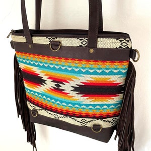 Könnte beinhalten: Eine braune Ledertasche mit einem bunten geometrischen Muster und Fransen. Die Tasche hat einen Reißverschluss und zwei braune Ledergriffe.