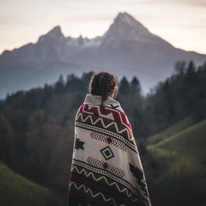 Op de afbeelding: Een persoon draagt een deken met patronen met rode, witte en bruine geometrische ontwerpen, voor een bergketen. De deken is over de schouders gedrapeerd en biedt warmte tegen de koele berglucht.