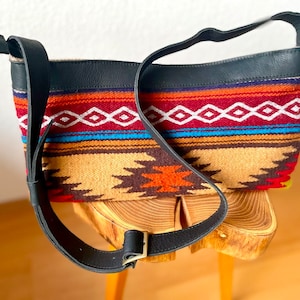 Peut inclure: Un petit sac tissé coloré avec une bandoulière en cuir noir. Le sac présente un motif géométrique dans des tons de rouge, orange, marron, bleu et blanc.