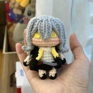 May include: A handmade crochet doll with gray hair, yellow eyes, and a black and yellow outfit. The doll is held in a person's hand. The doll's features include a white top, black skirt, and black shoes. The doll is a unique, handcrafted item.