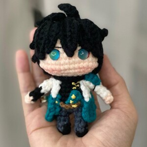 以下が含まれることがあります： 黒い髪とターコイズ色の目を持つ手作りのかぎ針編み人形。人形は、金色のアクセントが付いたターコイズと白の衣装を着ています。人形は人の手で持たれています。