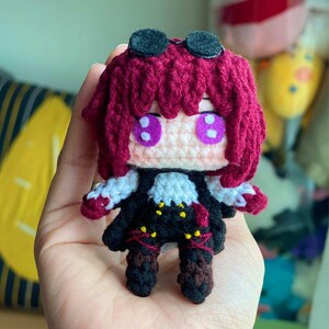 Op de afbeelding: Een handgemaakte gehaakte pop met een anime-geïnspireerd ontwerp. De pop heeft fel magenta haar, grote paarse ogen en een zwart-witte outfit. Het heeft een kleine, ronde, zwarte vilten bril en wordt in een hand gehouden.