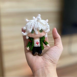 Op de afbeelding: Een kleine, handgemaakte gehaakte pop met wit haar, groene ogen en een groene jas. De pop heeft een wit shirt met een rood ontwerp, een zwarte broek en bruine schoenen. De pop wordt in een hand gehouden.