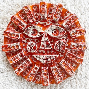 Puede incluir: Un rostro de sol redondo de cerámica naranja con detalles blancos. El sol tiene ojos, nariz y boca sonriente detallados. Los rayos del sol son rectangulares y texturizados, creando una pieza decorativa única.