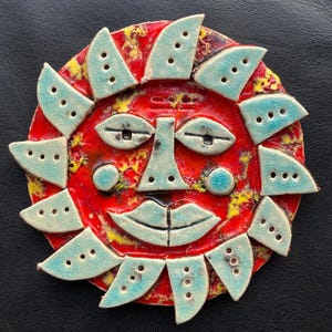 May include: A ceramic sun face with a red background and turquoise accents. The sun has a smiling face with closed eyes and triangular rays around the perimeter. The rays have three holes each.