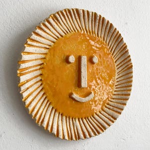 May include: A ceramic sun-shaped wall hanging with a smiling face. The sun has a bright yellow centre and a textured, off-white outer ring with radiating lines. The face features two small circles for eyes, a rectangular nose, and a curved mouth.