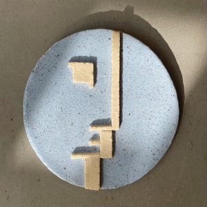 Puede incluir: Un plato circular de cerámica azul claro, moteado, con un diseño geométrico beige. El diseño consiste en una línea vertical y varias formas rectangulares. El plato está sobre una superficie gris.