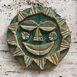 May include: A handmade ceramic sun face with a smiling expression. The sun has a turquoise glaze with tan accents, and triangular rays around the perimeter. The face features geometric shapes for the eyes, nose, and mouth.