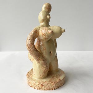May include: A ceramic sculpture of a nude figure on a rounded base. The sculpture is mainly beige with reddish-brown speckling. The figure has prominent breasts and an arm wrapped around the body.