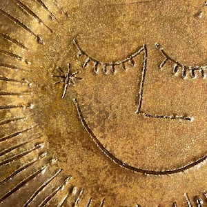May include: Close-up of a gold-coloured, textured ceramic plate with a sun-like design. The sun features a smiling face with closed eyes and a star-like element. The plate has a crackled surface.