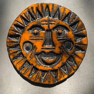 May include: A ceramic sun-shaped wall hanging with an orange and black glaze. The sun has a smiling face with circular eyes, a triangular nose, and a wide, smiling mouth. The sun's rays are triangular and point outwards.