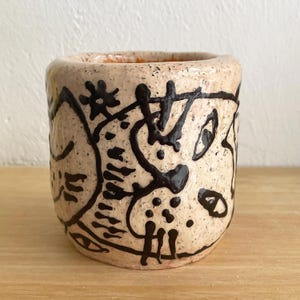 May include: A handmade ceramic cup with a beige speckled finish. The cup is adorned with black abstract line art. The interior of the cup is a light orange color. The cup sits on a wooden surface.