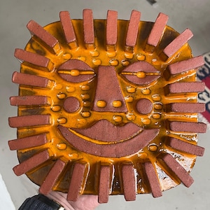 May include: A ceramic sun-shaped dish with a smiling face. The dish has an orange glaze with a brown face and brick-like rays. The design includes oval eyes, a nose, and a smiling mouth, creating a cheerful, decorative piece.
