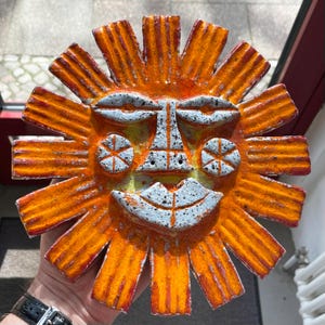 May include: A ceramic sun face sculpture with a smiling expression. The sun has orange rays with red edges, a white face with gray speckles, and yellow accents. The sculpture is handmade and decorative.