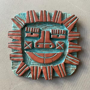 May include: A square, ceramic wall hanging featuring a stylized face. The face has a turquoise background with a smiling mouth, nose, and eyes in a reddish-brown color. The sun-like rays around the face are also reddish-brown.