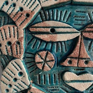 May include: A close-up of a ceramic wall hanging with a blue, white and brown colour scheme. The design features geometric shapes and patterns, including a large eye shape.