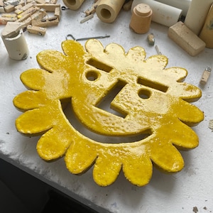 Könnte beinhalten: Ein hellgelbes, sonnenförmiges Keramikobjekt mit einem lächelnden Gesicht. Die Sonne hat blütenblattartige Ränder und ausgeschnittene Augen und Mund. Das Objekt befindet sich auf einer hellen Oberfläche, im Hintergrund befinden sich weitere Keramikteile.