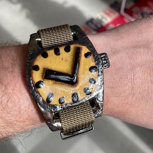 May include: A handcrafted watch with a distinctive, rustic aesthetic. The watch face is yellow with black markings and hands, set in a dark, textured metal casing. It has a brown fabric strap with a metal clasp, creating a unique, artistic accessory.