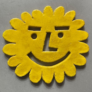 May include: A yellow ceramic sun-shaped coaster with a smiling face. The sun has a face with rectangular eyes, round pupils, and a nose. The sun's rays are petal-shaped. The coaster is a bright, cheerful yellow.