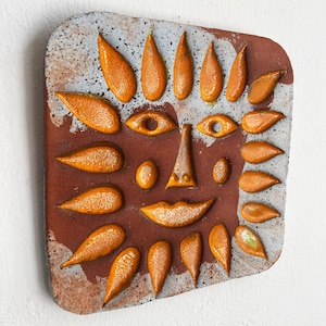 May include: A square ceramic wall art piece with a textured surface, depicting a stylized face. The face is made up of orange teardrop shapes, eyes, a nose, and a smiling mouth. The background is a mix of red and grey.