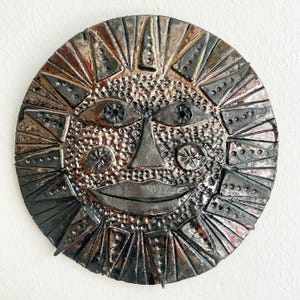 May include: A circular ceramic sun face sculpture with a textured surface. The sun features a smiling mouth, triangular nose, and stylized eyes. The sun's rays are made of triangles and the colour palette includes shades of brown, grey, and black.