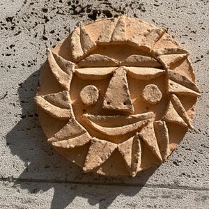 May include: A terracotta sun face sculpture with a smiling expression. The sun has triangular rays around the perimeter and a detailed face with a triangular nose, circular eyes, and a curved mouth. The sculpture is mounted on a textured, light-coloured wall.