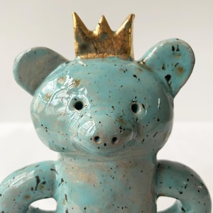 May include: A ceramic figurine of a blue bear wearing a gold crown. The bear has a speckled blue glaze and a shiny finish.
