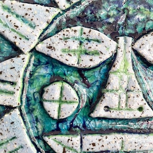 May include: Close-up of a ceramic artwork with a face-like design. The piece features white, green, and blue colours. The design includes shapes resembling eyes, a nose, and a mouth, with a textured, crackled glaze.