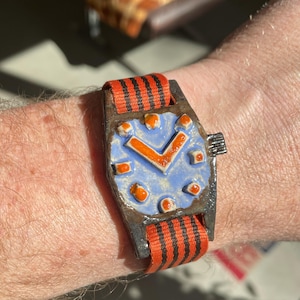 May include: A handmade watch with a blue face, orange hands and markers, and a black and orange striped strap. The watch has a rectangular case and a textured appearance. The watch is worn on a wrist.