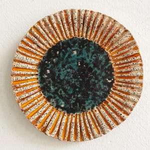 May include: A round ceramic plate with a textured surface. The centre is dark teal, surrounded by a black speckled area. The outer rim features vertical orange stripes and a speckled white background.