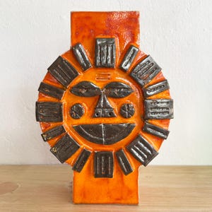 May include: A ceramic sun-shaped wall hanging with a smiling face. The sun has an orange background with dark brown rectangular rays and facial features. The piece is rectangular with a textured surface.