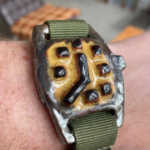 May include: A distinctive wristwatch featuring a square face and a green nylon strap. The watch face has a yellow background with black square hour markers and black hands. The watch case has a textured, metallic appearance.