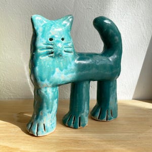 May include: A turquoise ceramic cat sculpture with a whimsical design. The cat has a long body, upright tail, and stylized paws. The sculpture is glazed with a glossy finish and sits on a wooden surface.