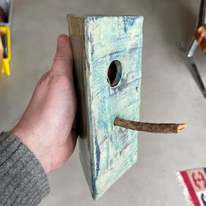 May include: A rectangular birdhouse with a light blue and green glazed finish. It has a circular entrance hole and a wooden perch. The birdhouse is held by a person.