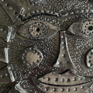 May include: Close-up of a metallic sculpture of a stylized face. The artwork has detailed textures, rivets, and circular patterns for eyes and cheeks. The overall colour is a shiny silver, with a slightly rough surface.