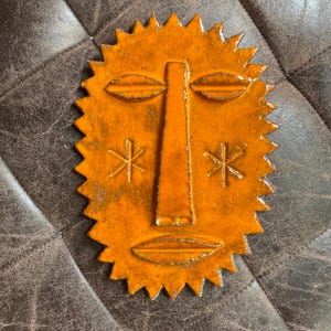 Könnte beinhalten: Eine glänzend orangefarbene Keramik-Wandplatte in Form eines stilisierten Sonnengesichts mit langer zentraler Nase, mandelförmigen Augen, horizontalem Mund und zwei sechsstrahligen Sternsymbolen auf den Wangen mit gezacktem Rand.