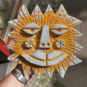 May include: A sun-shaped ceramic wall plaque featuring a textured white surface with black speckles and vibrant orange accents. The design includes a central face with almond-shaped eyes, a triangular nose, and a wide, smiling mouth.