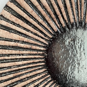 May include: Close-up of a ceramic plate with a sunburst design. The plate has a textured, light brown surface with radiating black lines. The centre of the plate is a darker, textured brown.