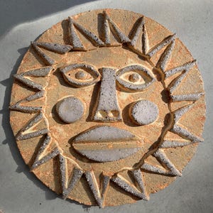 May include: A circular ceramic wall plaque featuring a stylised sun face. The piece has a textured, speckled light brown base with raised, grey-toned features including triangular sun rays, almond-shaped eyes, circular cheeks, and lips.