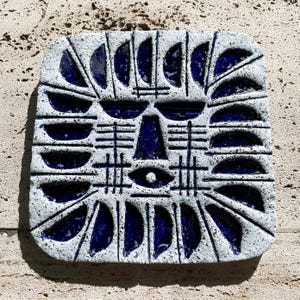 May include: Square ceramic tile with a textured surface and a face design. The tile has a white background with dark blue accents, including crescent shapes and geometric patterns. The design includes an eye and a nose.