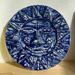 May include: A circular, blue ceramic sun face with a smiling expression. The sun's rays are triangular and surround the face. The face and rays have a textured, mottled appearance with white accents.
